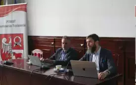 Pomieszczenie; przy stole siedzi dw&oacute;ch mężczyzn w marynarkach. Przed każdym z nich znajdują się laptopy oraz mikrofony. Z prawej strony znajduje się roll-up z napisem POWIAT Czarnkowsko-Trzcianecki oraz herbem powiatu.