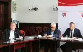 Pomieszczenie; przy stołach ustawionych w podkowę, siedzi trzech mężczyzn w marynarkach. Przed każdym z nich znajduje się mikrofon.