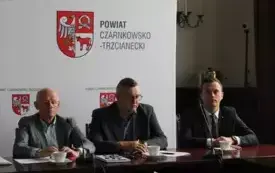 Pomieszczenie; przy stole siedzi trzech mężczyzn w marynarkach, przed każdym z nich znajduje się mikrofon oraz filiżanki na spodkach.