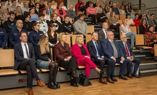 Pomieszczenie; na widowni siedzi grupka ludzi, słuchająca konferencji. W pierwszym rzędzie 3 kobiety, 4 mężczyzn.