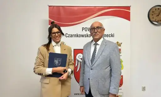 Pomieszczenie, na pierwszym planie kobieta i mężczyzna, oboje w okularach. Kobieta trzyma w ręku teczkę. Za nimi napis Powiat Czarnkowsko - Trzcianecki.