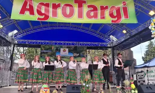 Scena, na niej ubrane w ludowe stroje kobiety i mężczyźni. Nad nimi napis: AgroTargi.