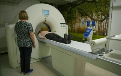 Pomieszczenie, na pierwszym planie kobieta badająca się tomografem, obok stoi kobieta przyglądając się badaniu.