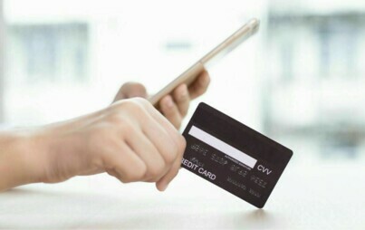 Na pierwszym planie dwie ręce, w jednej telefon komórkowy, w drugiej karta bankomatowa debetowa.